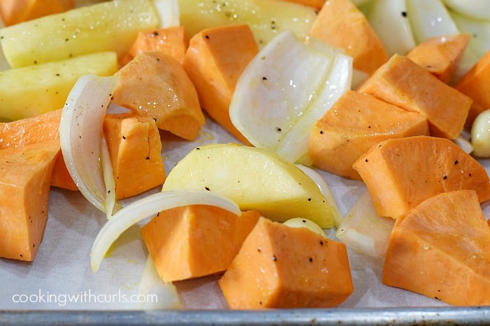 Wedges of sweet potato, onion, apples, and garlic cloves coated with oil and seasonings on a baking sheet.