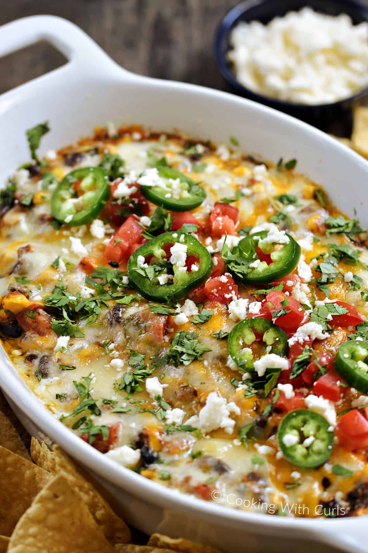 An oval baking dish filled with black bean dip topped with melted cheeses, crumbled queso, sliced jalapenos, diced tomatoes, and chopped cilantro surrounded by tortilla chips. 