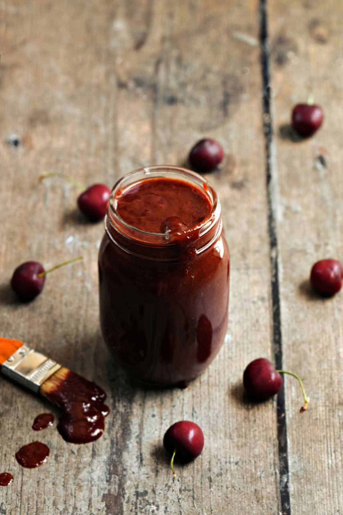 A jar of cherry chipotle bbq sauce surrounded by fresh cherries.