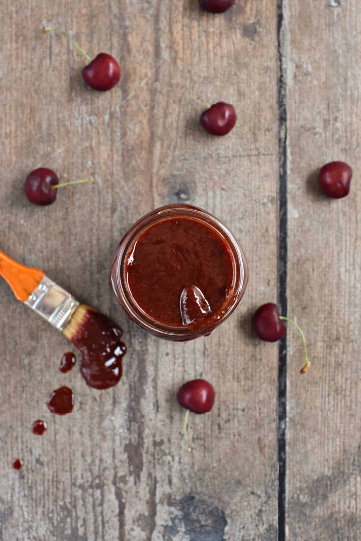 Cherry chipotle bbq sauce in a mason jar surrounded by fresh cherries and spilled sauce.