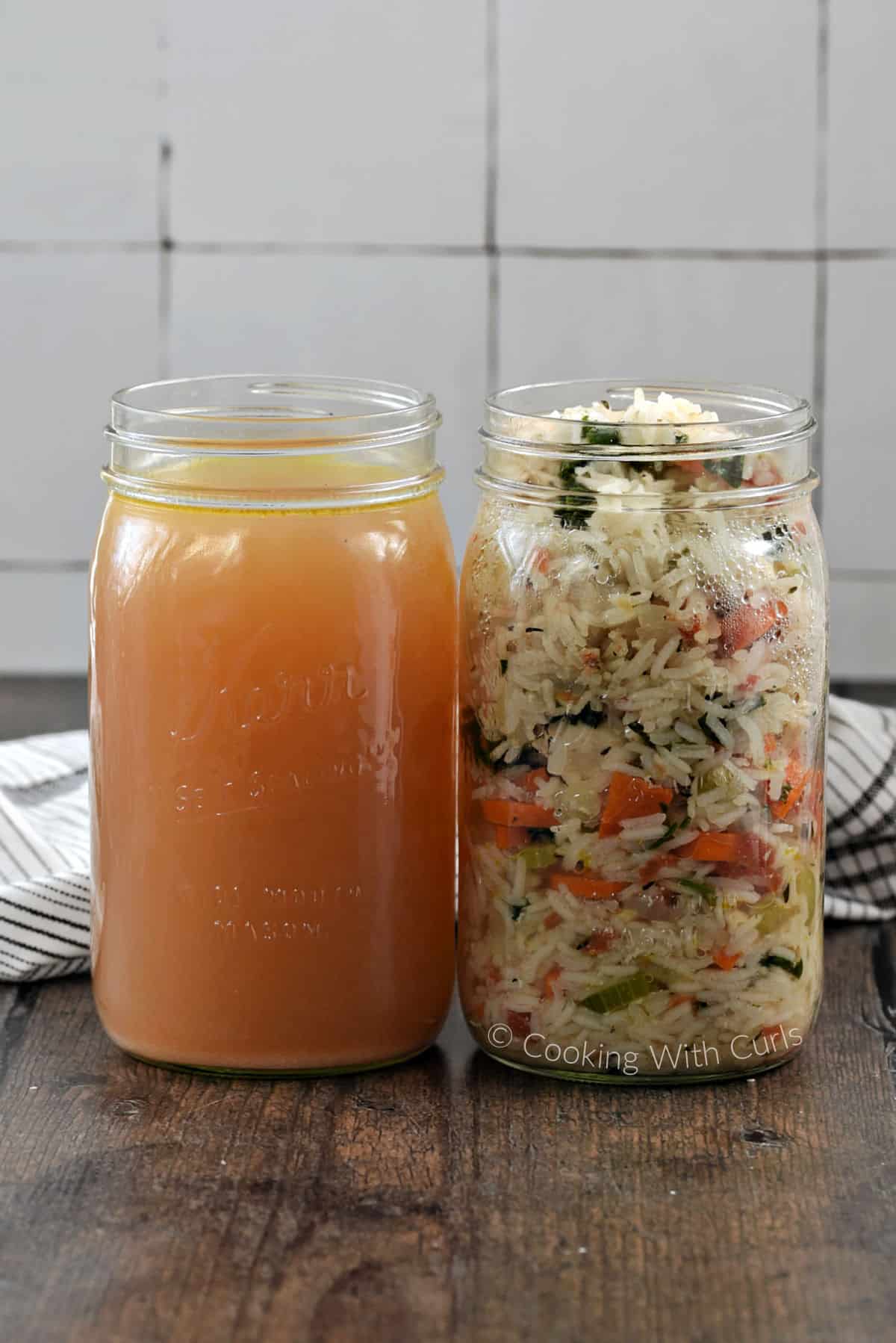 A jar of chicken stock and a jar of rice, carrots, kale, tomato, and celery sitting on a wooden surface. 