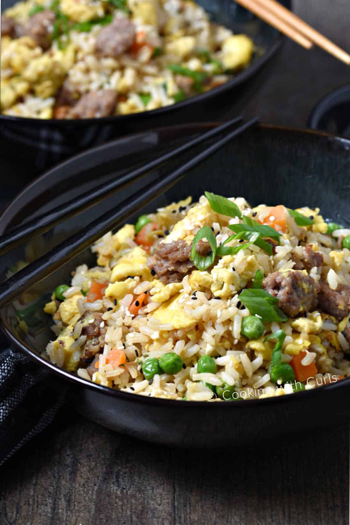 Fried rice, sausage, scrambled eggs, green onions, peas and carrots in a bowl with chopsticks on the edge. 