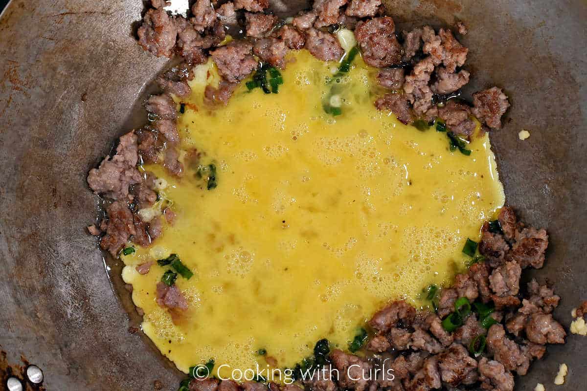 Raw eggs in the center of a wok with cooked sausage and green onions. 