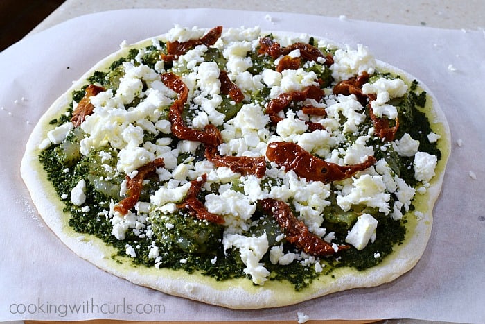 Raw pizza dough circle topped with pesto, shrimp, crumbled cheese, and slices of sun-dried tomatoes.