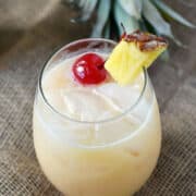 A creamy cocktail in an ice filled glass garnished with a pineapple wedge and cherry.