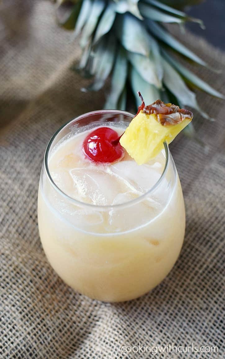 A creamy cocktail in an ice filled glass garnished with a pineapple wedge and cherry.