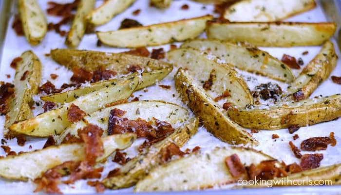Baked potato wedges topped with grated cheese, bacon, and herbs on a parchment lined baking sheet.