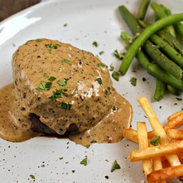 Creamy peppercorn sauce over a filet mignon on a plate with green beans and french fries.