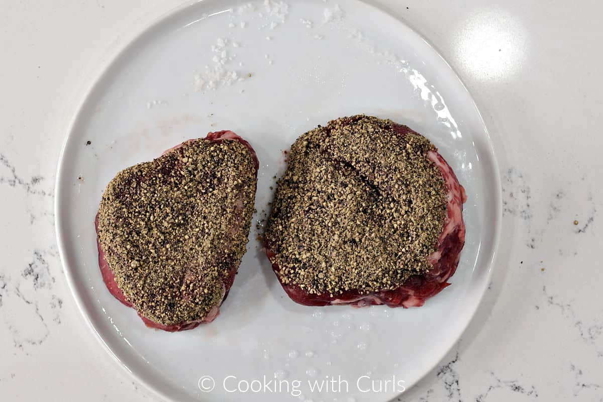 Two filet mignon coated with crushed black pepper on a plate. 
