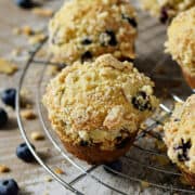 Five streusel topped blueberry muffins on a round wire cooling rack surrounded by fresh blueberries.