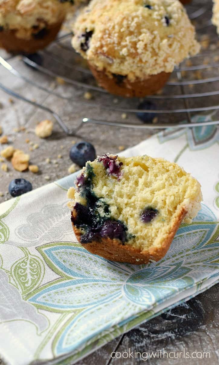 Blueberry muffin split in half laying on a fabric napkin with remaining muffins on a wire cooling rack in the background.