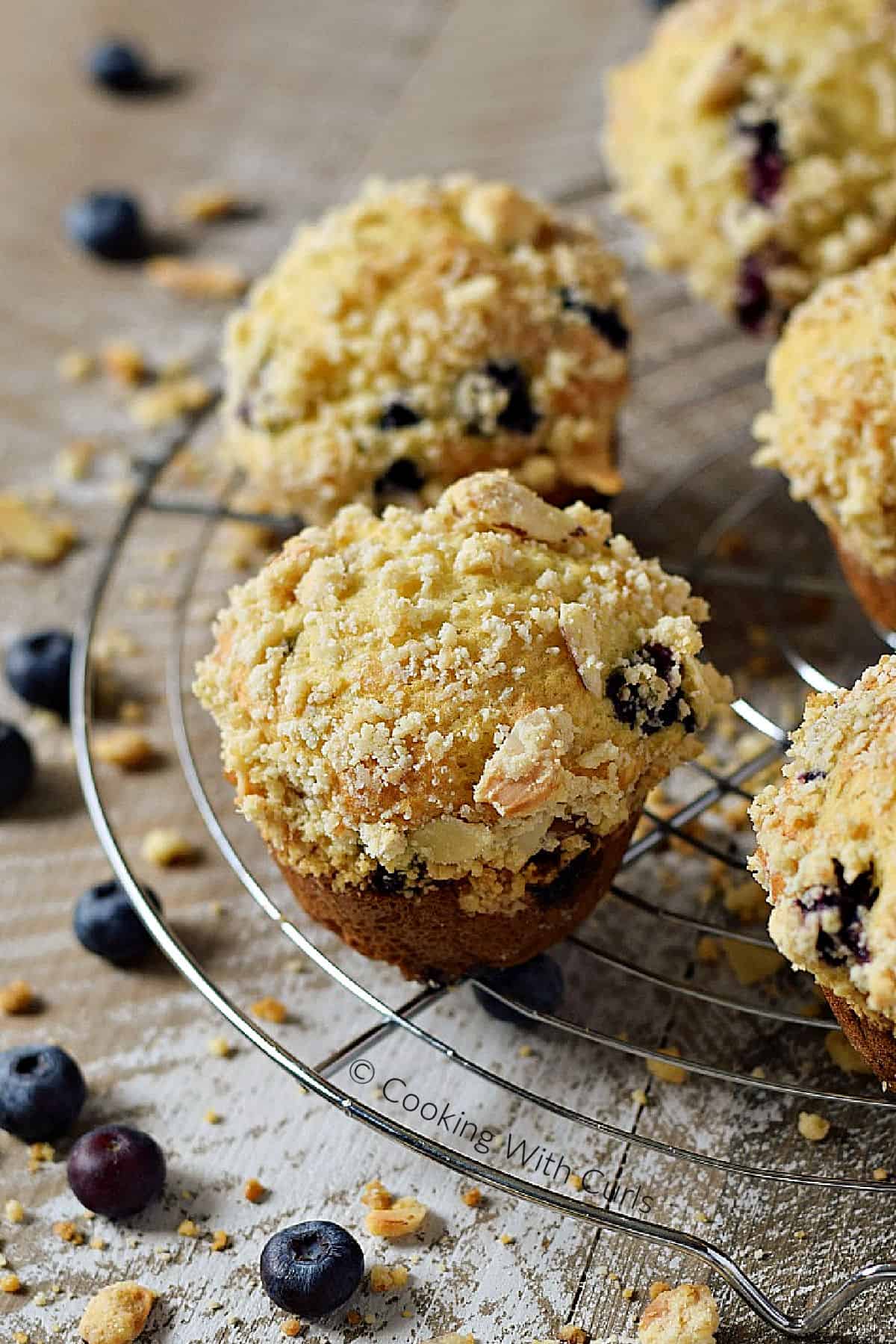 Five streusel topped blueberry muffins on a round wire cooling rack surrounded by fresh blueberries.