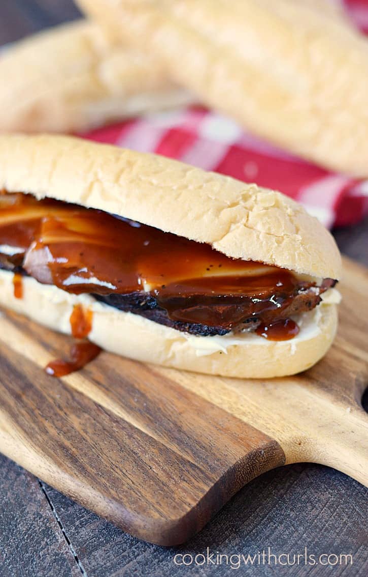 sandwich roll filled with sliced tri-tip and provolone cheese covered with barbecue sauce and sitting on a wooden cutting board with extra rolls in the background