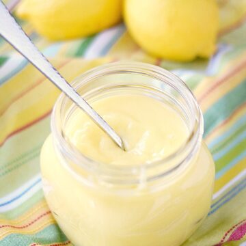 lemon curd in a large glass jar with a spoon inside and two lemons sitting on a striped napkin in the background