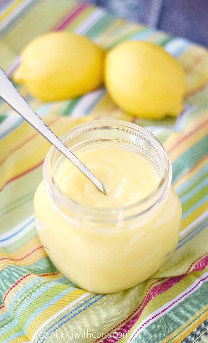 lemon curd in a large glass jar with a spoon inside and two lemons sitting on a striped napkin in the background