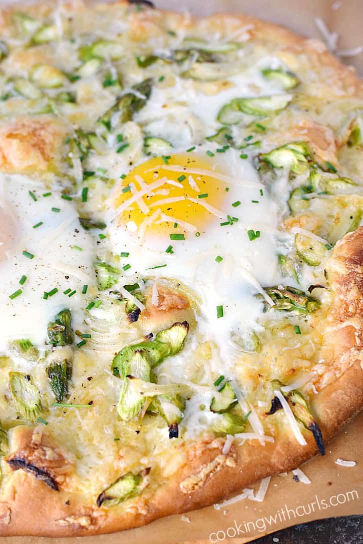 close-up image of a pizza topped with eggs, thinly sliced asparagus, cheese and chopped parsley