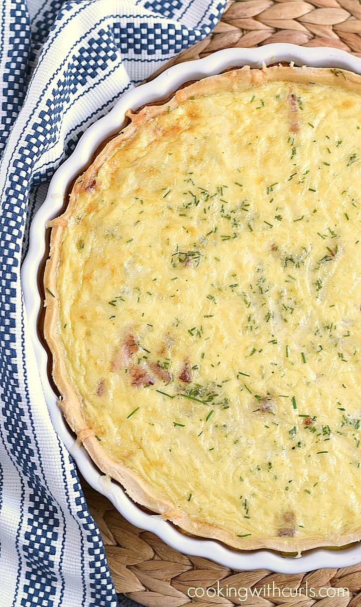Classic Quiche Lorraine in a ceramic white tart pan sitting on a woven place mat and and blue and white stripped napkin