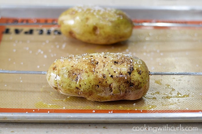 Two raw potatoes rubbed with oil and salt on a silicone lined baking sheet with a metal skewer through one of the potatoes.