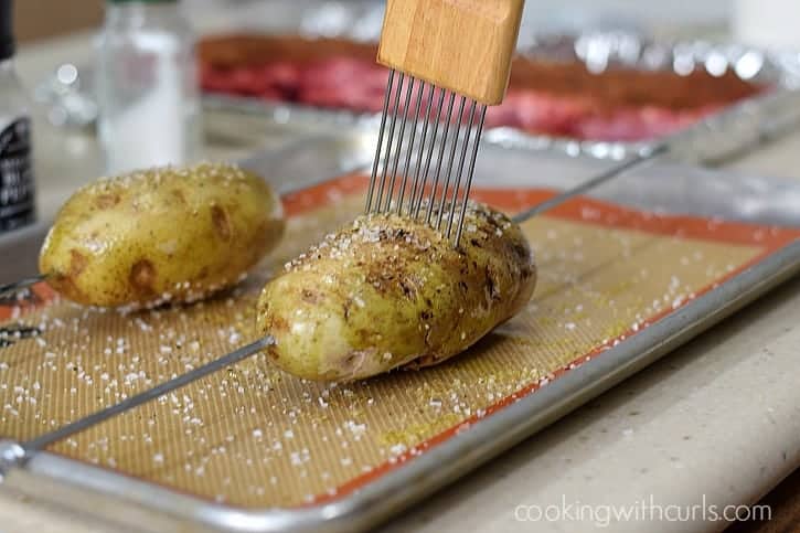Two skewered potatoes on a baking sheet getting vent holes poked into the top.