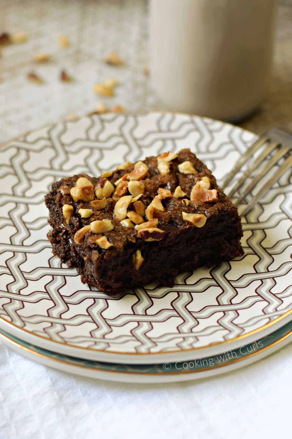 A hazelnut brownie on a stack of plates with a fork on the edge and glass of milk in the background.