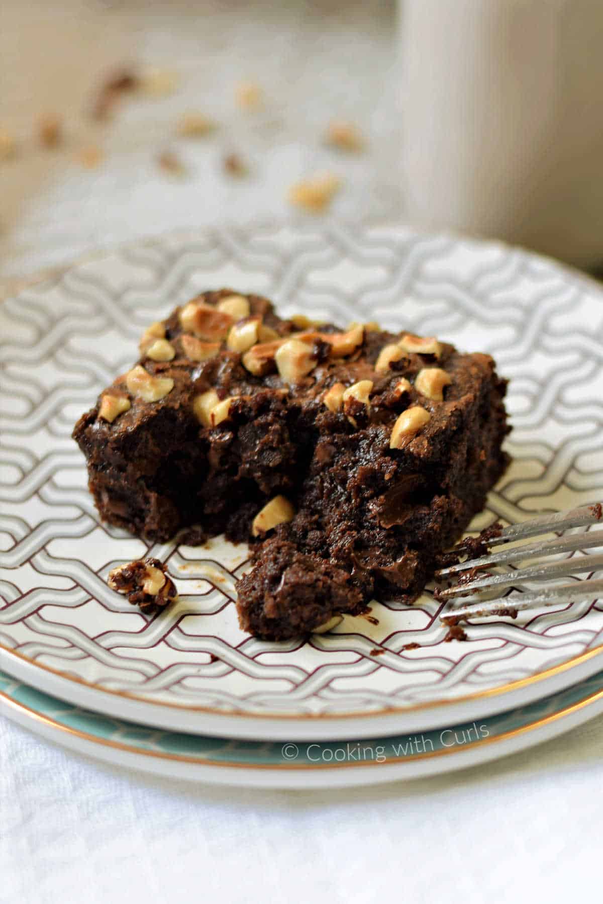 A single hazelnut brownies on a plate with a fork and glass of milk in the background.