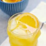 a glass of orange peach mango spritzer sitting on a blue striped napkin with a blue bowl of orange slices on the left side