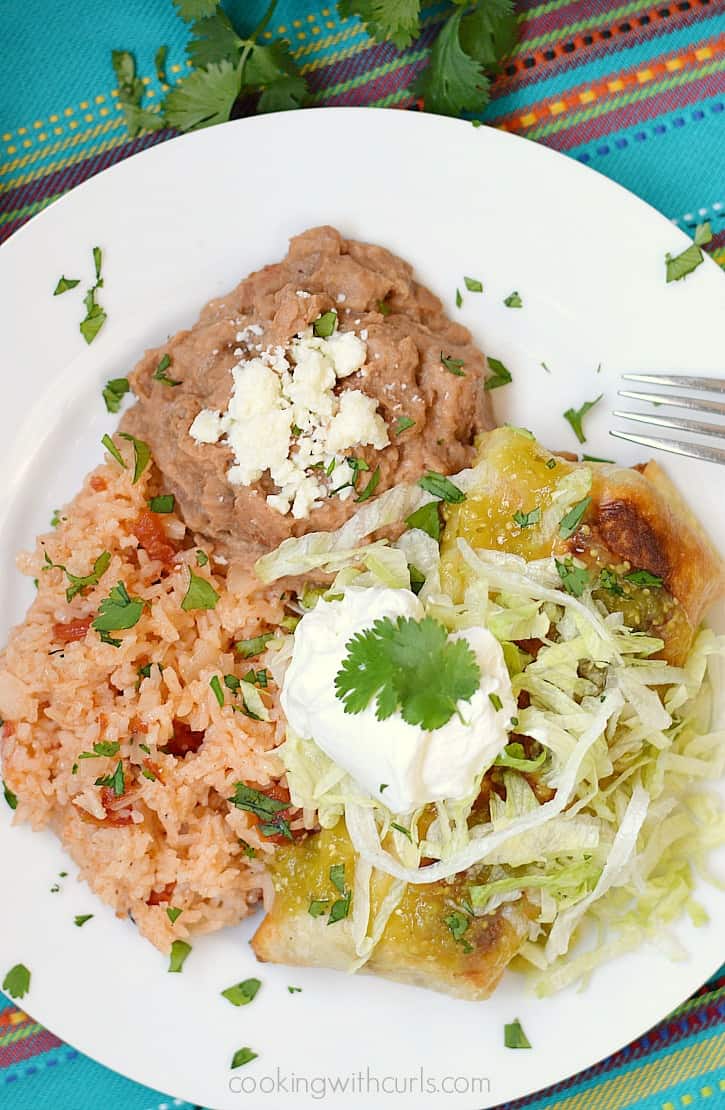 Looking down on a plate with Shredded Beef Chimichangas topped with salsa verde, shredded lettuce, and sour cream next to Spanish rice and refried beans.