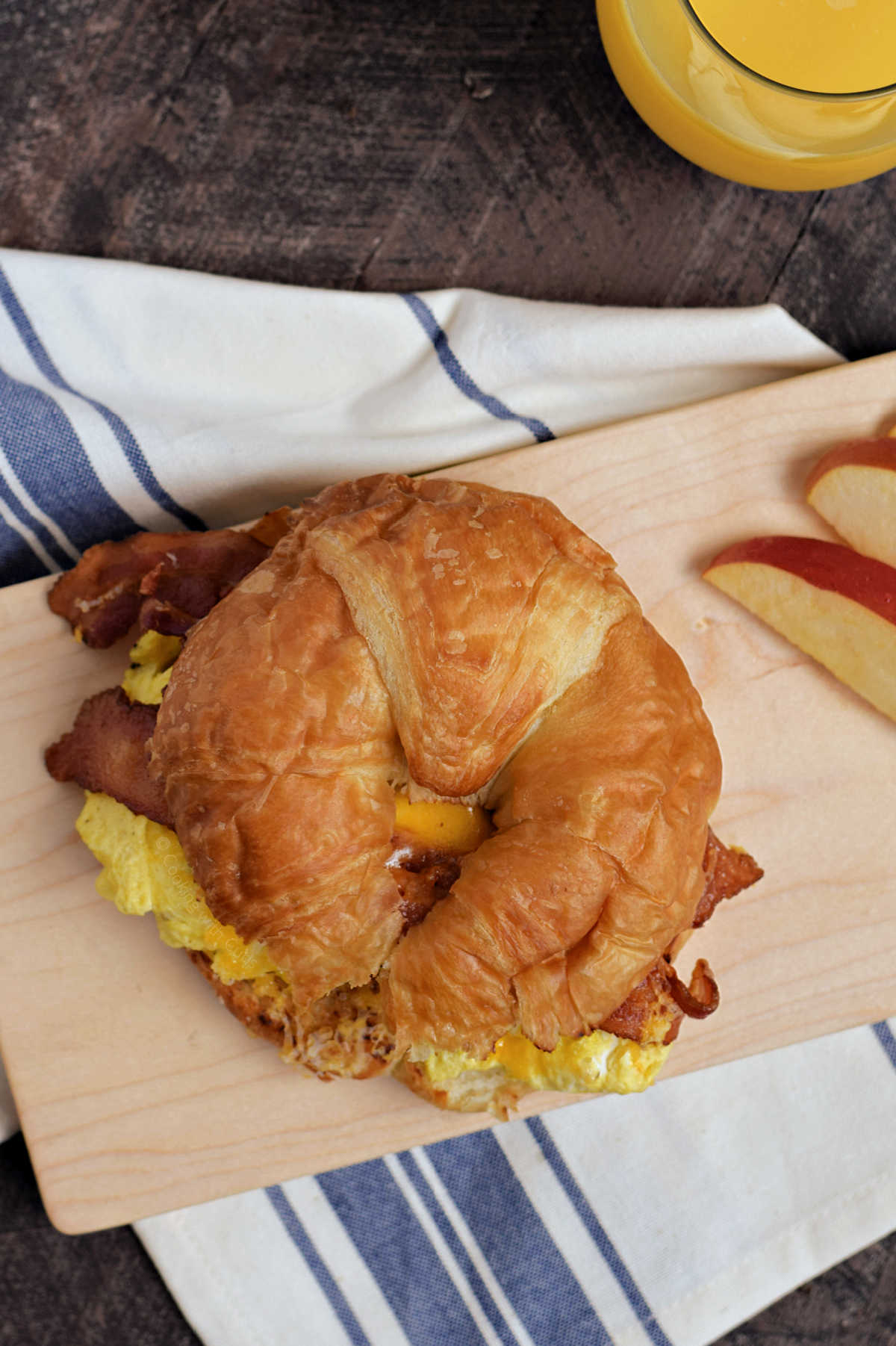 Bacon, scrambled egg, and melted cheese on a croissant with orange juice and apple slices in the background.