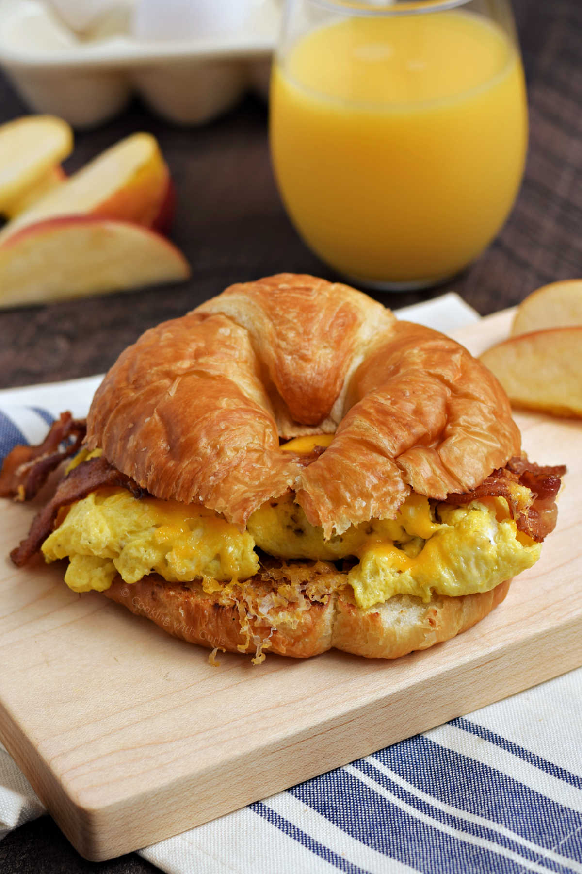 Bacon, scrambled egg, and melted cheese on a croissant sitting on a wooden board, with orange juice in the background.