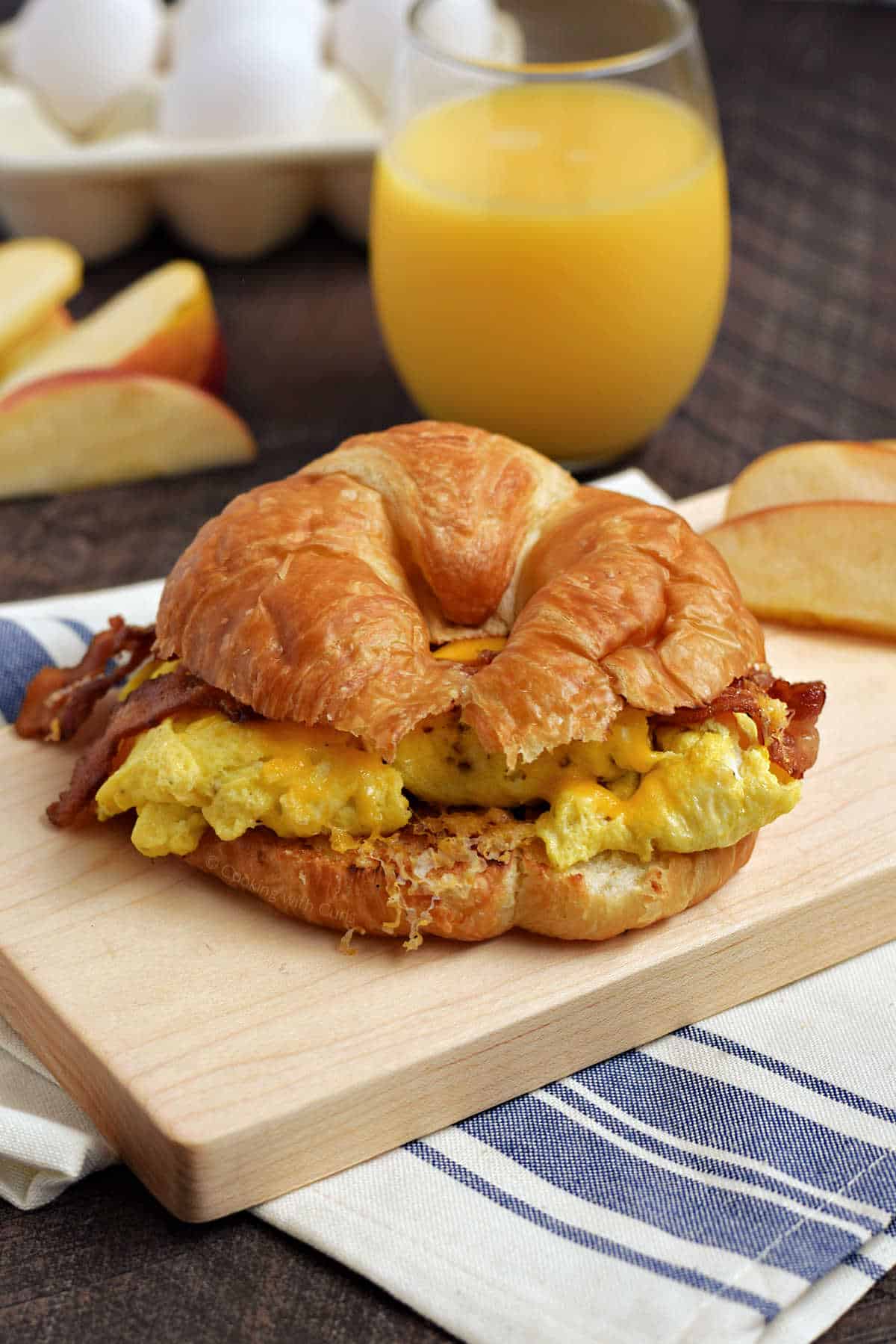 Bacon, scrambled egg, and melted cheese on a croissant with orange juice in the background.