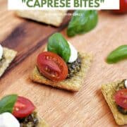Five crackers topped with basil pesto, mozzarella pearl, cherry tomato half, and basil leaf on a serving board with title graphic across the top.