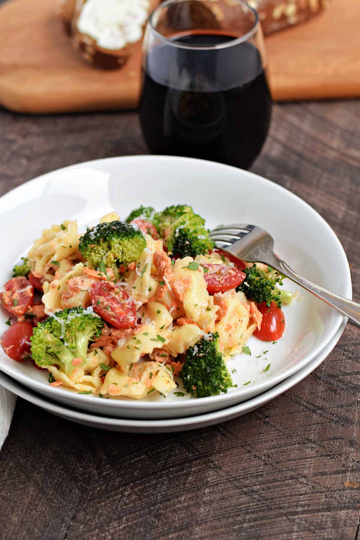 Cheese tortellini, salmon, broccoli, and cherry tomatoes in a bowl with a glass of red wine in the background. 