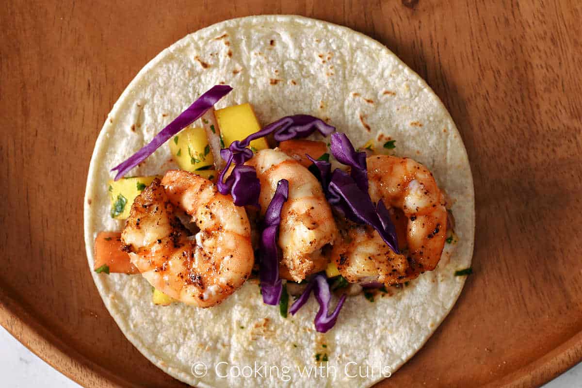 A corn tortilla topped with mango salsa, grilled shrimp, and shredded red cabbage on a wood plate.