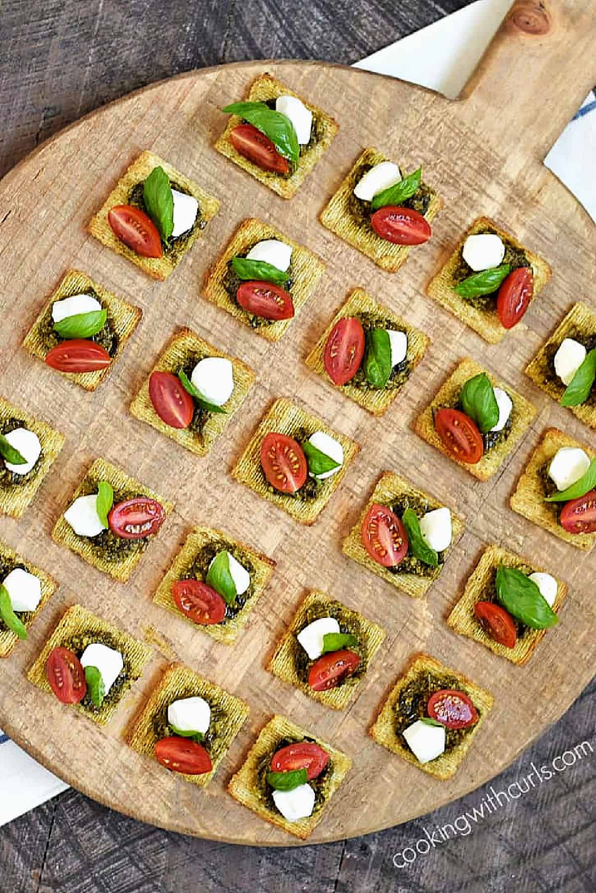 Twenty three crackers topped with basil pesto, mozzarella pearl, cherry tomato half, and basil leaf on a serving board.