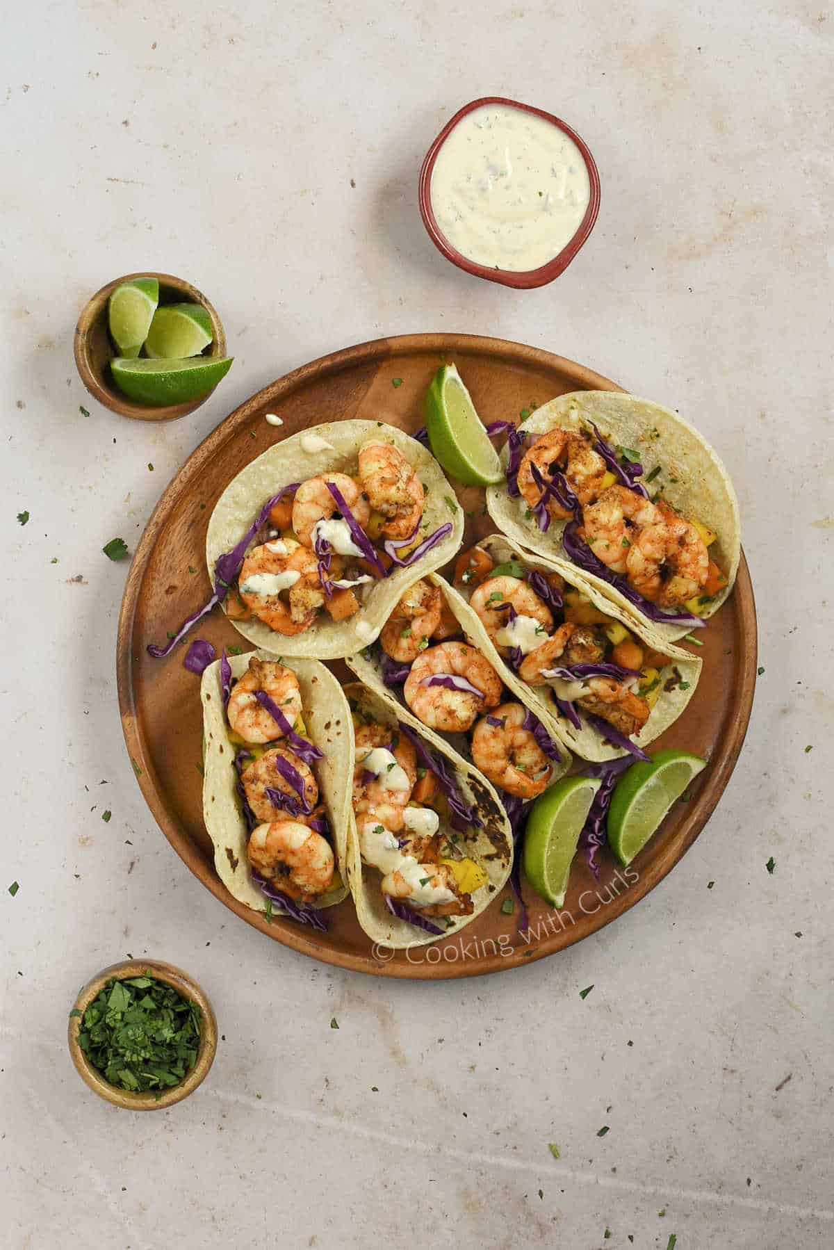 Looking down on a plate of grilled shrimp tacos with small bowls of lime wedges, chopped cilantro, and chipotle crema.