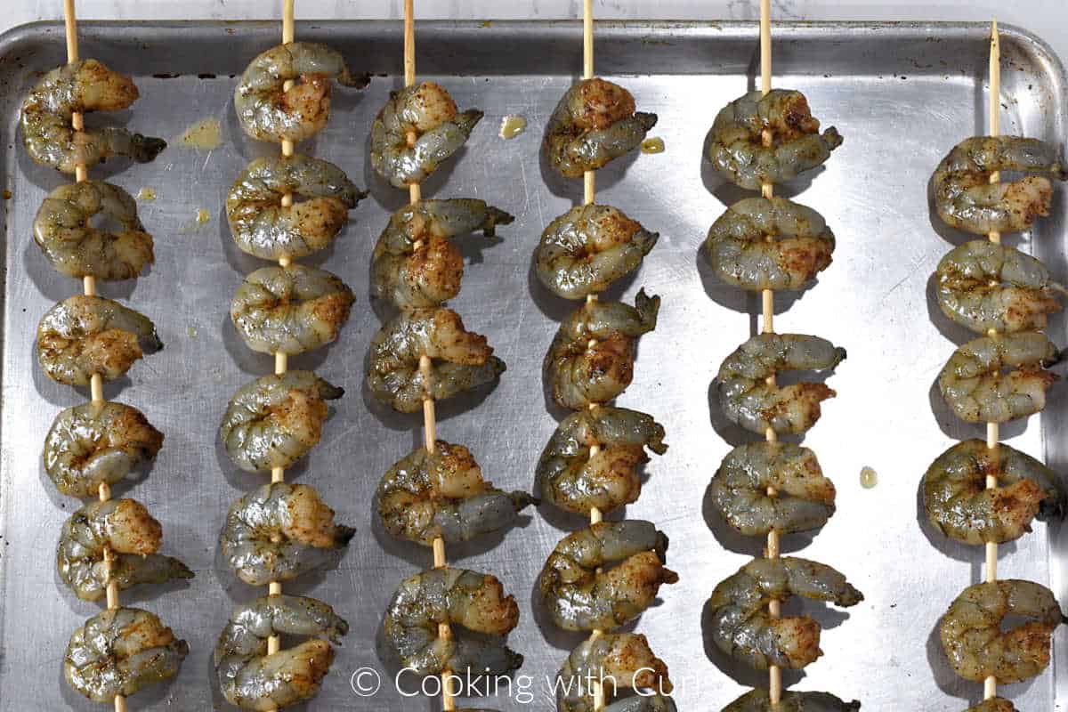 Six skewers with seasoned shrimp laying over a baking sheet.