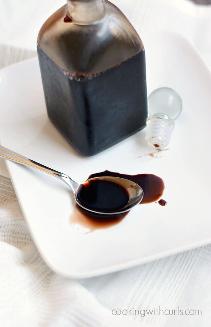 Balsamic Glaze in a square bottle sitting on a white plate with a spoon spilling additional glaze onto the plate.