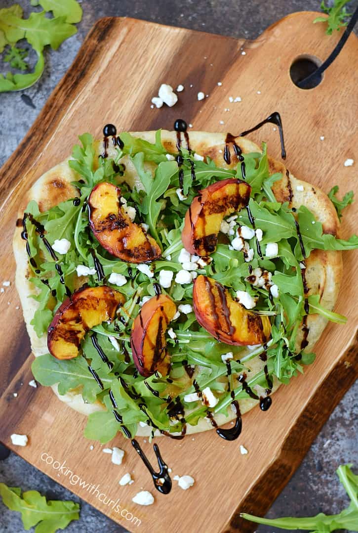 Looking down on a pizza topped with grilled peaches, arugula leaves, crumbled goat cheese and balsamic drizzle.