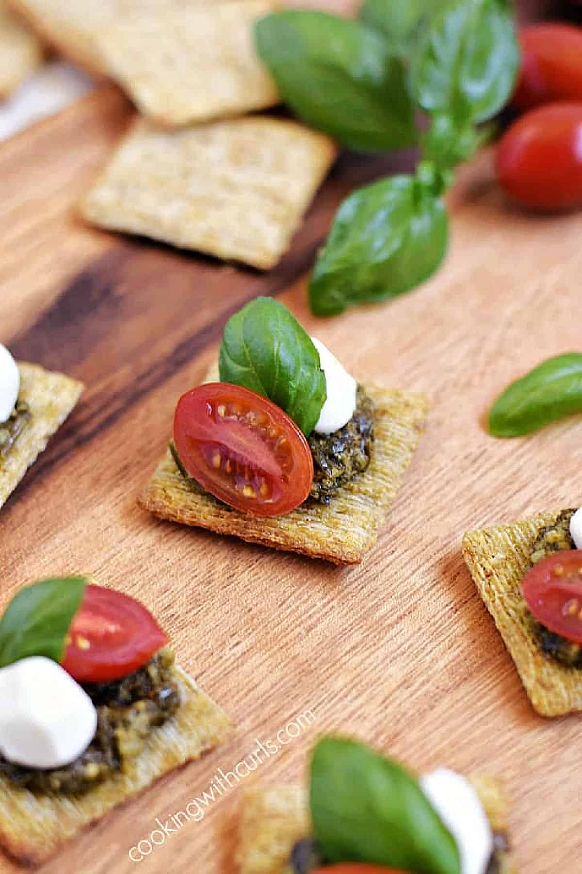 Five crackers topped with basil pesto, mozzarella pearl, cherry tomato half, and basil leaf on a serving board.