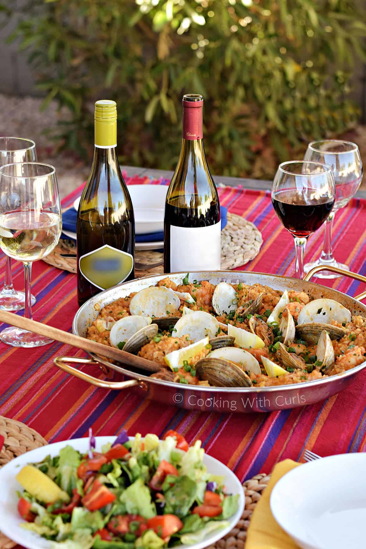 Seafood and rice in a large paella pan outside on a striped tablecloth surrounded by wine bottles and salad.