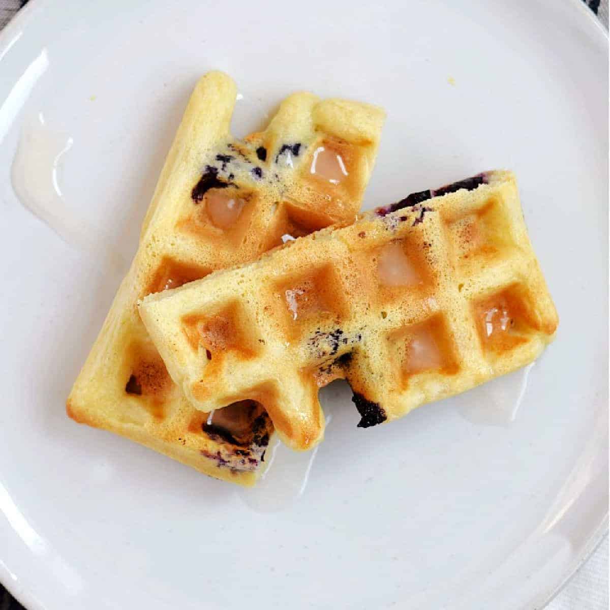 A blueberry and lemon waffle that is cut in half drizzled and with glaze on a white plate.