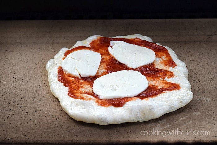 Pizza dough with tomato sauce and mozzarella cheese slices on a baking stone.