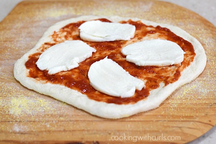 A circle of pizza dough topped with four slices of fresh mozzarella and pizza sauce on a cornmeal covered pizza peel.