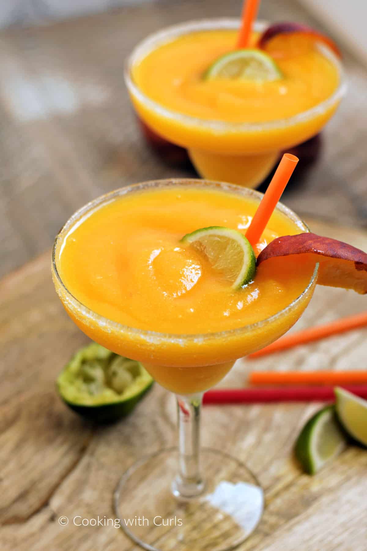 Two frozen peach margaritas in sugar rimmed glasses with a lime wedge and peach slice.