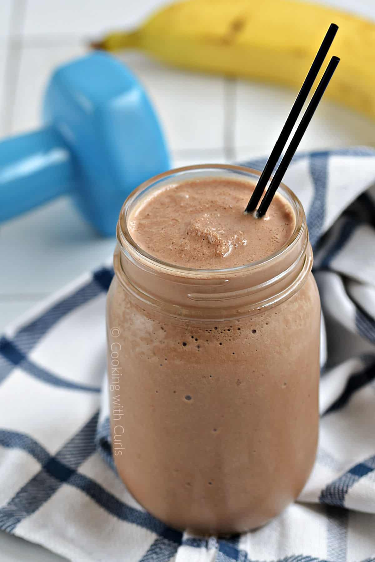 A chocolate smoothie in a mason jar with a dumbbell and banana in the background.