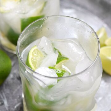 two glasses of ice and limes on a silver tray filled halfway with a caipirinha