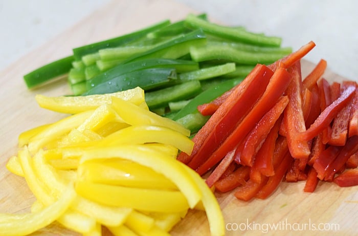 Thinly sliced green, red, and yellow bell peppers on a cutting board.