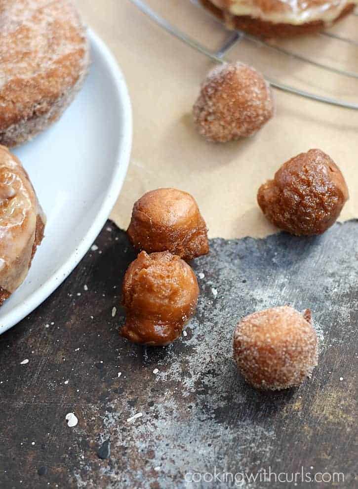 Three glazed and two sugar coated doughnut holes on a table next to a plate of full-size doughnuts.