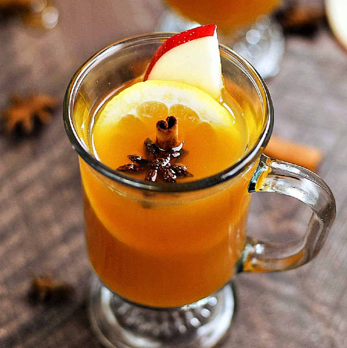 Whiskey cocktail in a glass footed mug garnished with lemon wheel, apple slice, cinnamon stick, and a star anise.