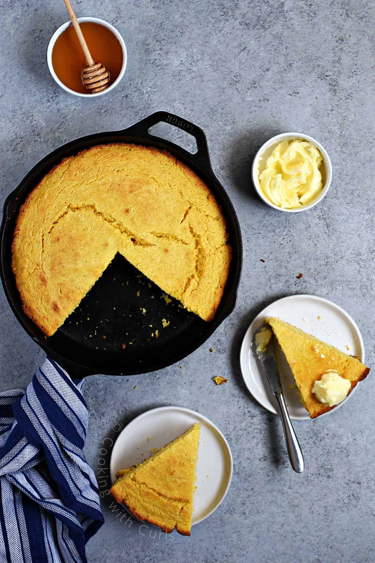Looking down on cornbread baked in a cast iron skillet with two slices cut out and placed on small plates topped with butter and honey.
